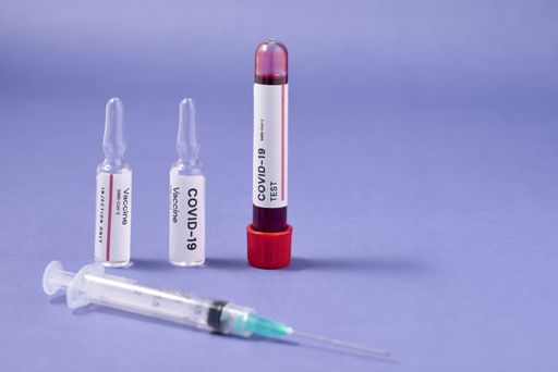 A closeup of a syringe needle with the COVID-19 test and vaccine ampoules on a purple surface