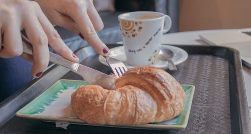 A selective focus shot of a female's hands cutting the croissant next to the coffee mug