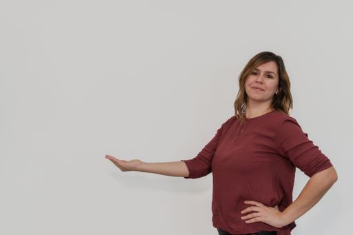 A Caucasian female stands against a white wall, her right arm outstretched and her palm facing outward