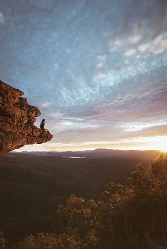 A vertical shot of a young girl sitting on the edge of a cliff and enjoying the mesmerizing sunset