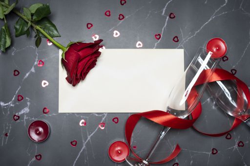 A top view of champagne glasses with a red ribbon, candles, small hearts, rose and white paper with copy space