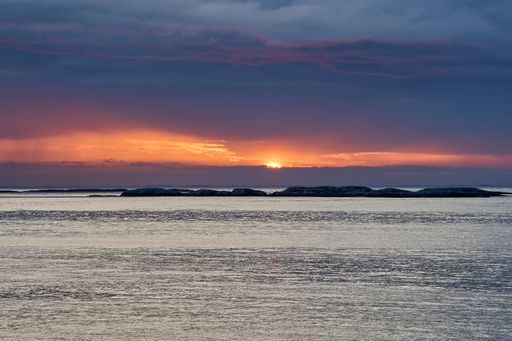 Dramatic sunset over the north Atlantic ocean at the rugged coastline of northern Norway, with an orange glowing sun and dark clouds.