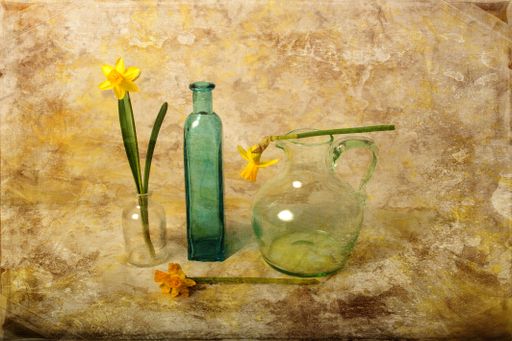 A still life with daffodils, a glass bottle, a pitcher, and a vase against a textured background.