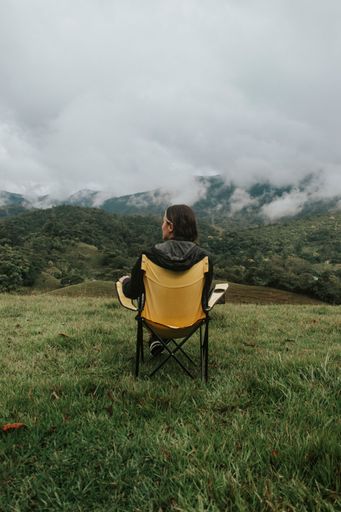 A woman sitting on a foldable chair in a grassy valley surrounded by green hills.