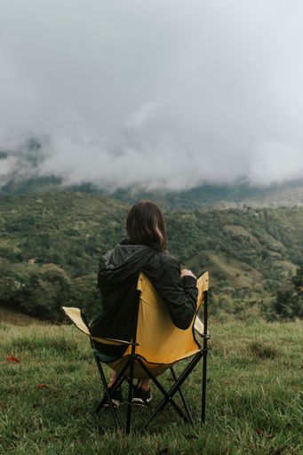 A woman sitting on a foldable chair in a grassy valley surrounded by green hills.