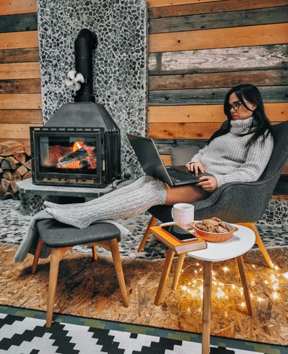 A Caucasian woman sitting comfortably in the chair next to the fireplace with her laptop on her knees