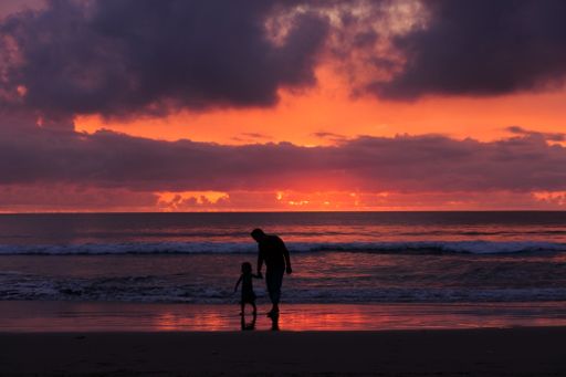 A male adult with a kid walking along the sandy shore near the waves on background of burning sunset