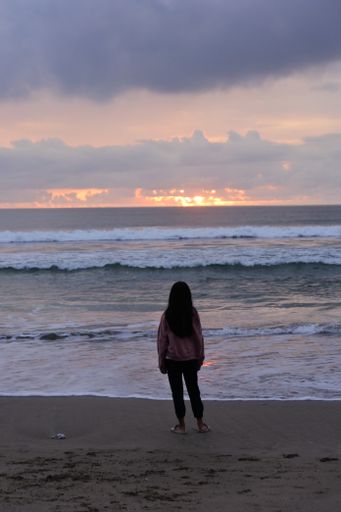 a vertical shot of a female shot from behind on the sandy shore near the waves at sunrise