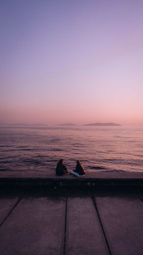 A vertical shot of two people sitting on a stone surface near the tranquil sea against a pink sky