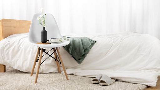 A closeup of a simple chair near a bed with a cup of coffee on it against a white wall
