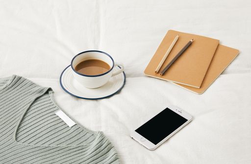 A high angle shot of a cup of coffee on bedsheets with sketchbooks, phone, and a sweater on it