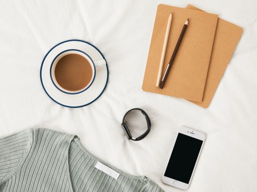 A top view of a cup of coffee on bedsheets with sketchbooks, phone, and a sweater on it