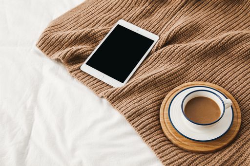 A high angle shot a tablet with a cup of coffee on a sweater on bedsheets