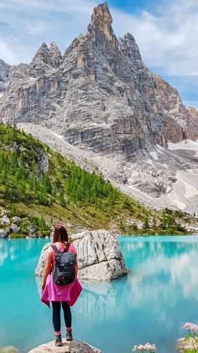 A vertical shot of a female with a backpack standing on a stone near a crystal clear lake