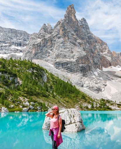 A vertical shot of a female with a backpack standing on a stone near a crystal clear lake