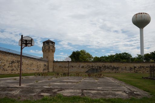 An old basketball court lies in front of an aging brick wall, which is situated in front of an ancient water tower