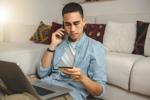 A young Hispanic male holding a credit card and talking on the phone for an error while making an online purchase on the laptop
