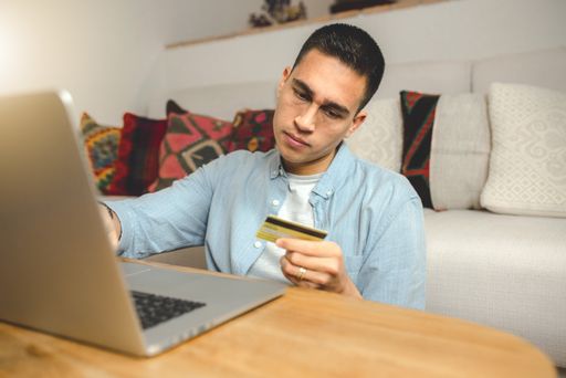 A shallow focus of a young Hispanic man holding a credit card for payment using a laptop computer at home