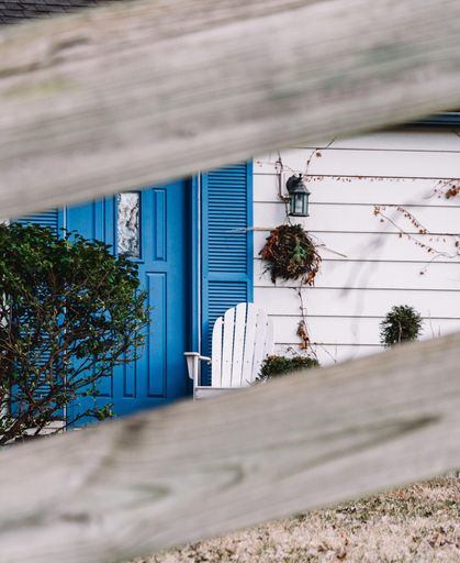 An exterior of a white house with a blue door shot through the wooden fence