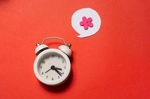 A high angle shot of an alarm clock, a pink flower in a cloud over it on a red surface