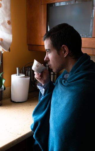 A vertical shot of a young man wrapped in a blanket drinkin coffee in the kitchen