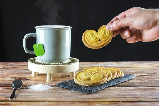 A hand of a person holding a puff pastry cookie next to a cup of hot tea