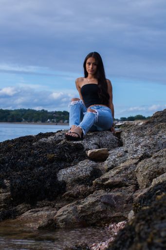 A vertical shot of an attractive Hispanic female wearing a strapless crop top sitting on sea rocks