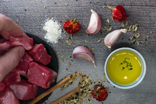 A top view of a male hand taking raw cut meat from a table with garlic cloves, dried pepper, oil