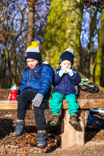 Two small kids wearing warm clothes eating and drinking on a wooden bench in the Debiec forest on a cold winter day.