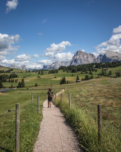 a vertical shot of a person walking on  a dirt path with the Plattkofel mountain in the background in Compatsch Italy