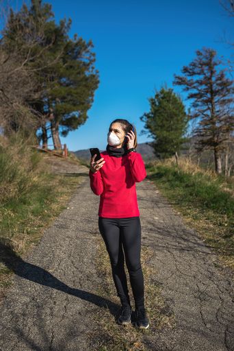 A vertical shot of a young Caucasian female in a medical mask listening to the music with headphones in a park on a sunny day