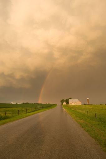 A vertical picture of a road surrounded by a field under a cloudy sky and a rainbow during the sunset