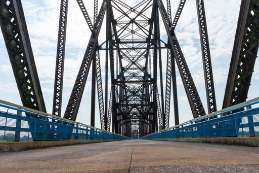 A low angle view of road and truss bridge above water and the forest in the background