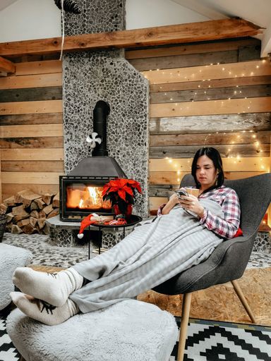 A Caucasian woman in sleepwear scrolling through her phone while drinking coffe by the fireplace at home