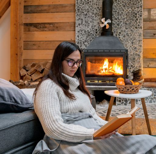 A Caucasian woman in sweater and eyeglasses reading book by the fireplace at home