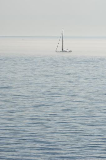 A vertical picture of the sea with a ship on it covered in the fog under a cloudy sky