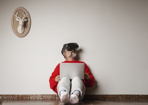 An adult male with VR glasses sitting on the floor and using his laptop