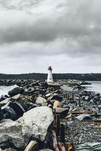 A vertical shot of a rocky shore with broken wooden road against with a lighthouse in the background