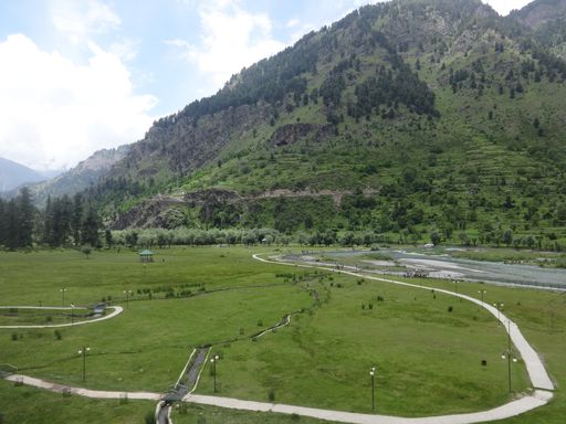 An aerial shot of a mountainous area with green trees and a green field with a narrow road and streetlights