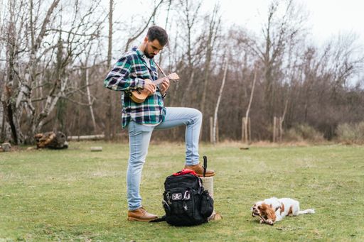 A Hispanic male playing a song on his ukulele in the woods with his dog laying on the grass