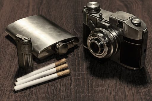 A selective focus shot of a vintage camera, a flask, cigarettes, and a cigarette lighter on a table