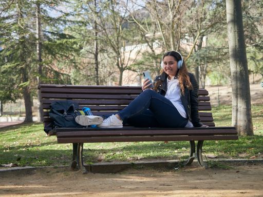 A young female with headphones using her smartphone while sitting on a wooden bench in the park