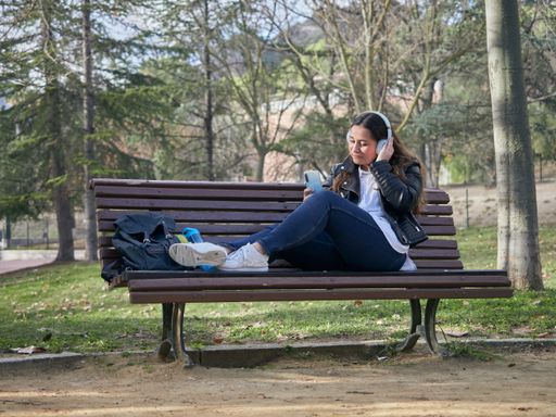 A young female with headphones using her smartphone while sitting on a wooden bench in the park