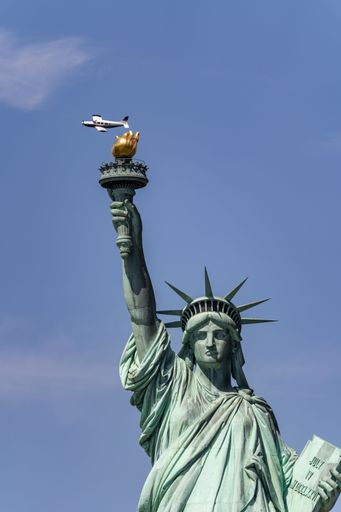The Statue of Liberty on a sunny day, with a small airplane flying in the blue sky in the background. Vertical shot