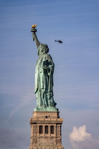 A vertical shot of the Statue of Liberty on a sunny day, with a helicopter flying in the blue sky in the background