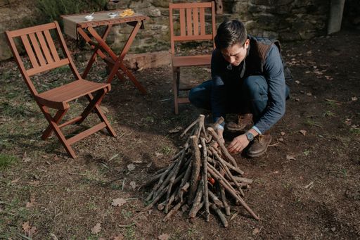 A young man preparing a pile of wood for fire with a table and chairs on the side - romantic dinner