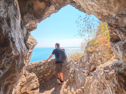 man with short pants, t-shirt, hiking backpack stands at a cliff viewpoint and watching the sea view panorama on the rilkeway in italy in summer