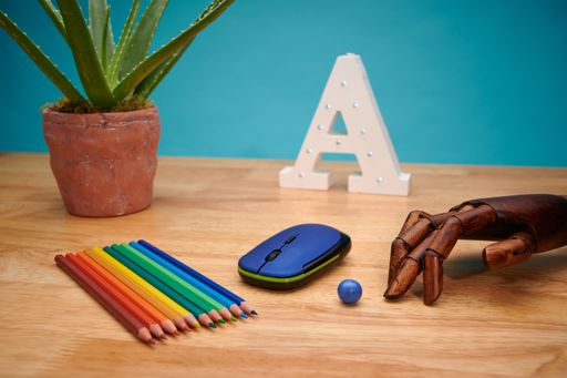 A selective focus shot of a computer mouse with multicolored pencils, articulated wooden hand, and aloe plant on a wooden table