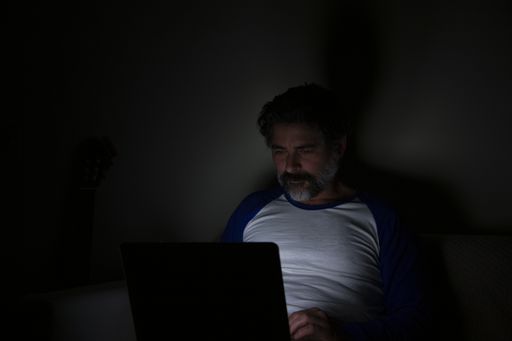 A middle-aged Caucasian bearded male sitting on a sofa at home at night, working on the laptop