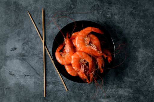 A top view of cooked shrimp on a black bowl with sticks on a gray ceramic surface